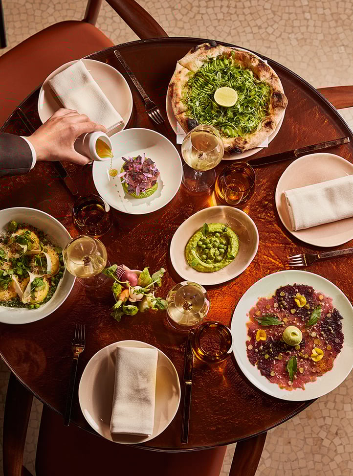 Overhead view of an elegant restaurant table set for three, featuring a vibrant selection of modern Mediterranean dishes including a green vegetable pizza with a lime wedge, avocado tartare topped with edible flowers, pea hummus, vegetable ravioli with herbs, fresh crudités, and beef carpaccio with microgreens and flower petals. A person pours dressing over a dish, and glasses of white wine are placed beside each plate. The table is copper-toned with soft lighting and upholstered chairs.
