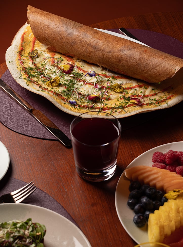 Close-up of a breakfast table featuring a large, rolled dosa topped with edible flowers, herbs, and sauce, placed on a dark placemat. Surrounding dishes include a plate of fresh fruit with raspberries, cantaloupe, blueberries, and pineapple, a green salad, and a glass of dark juice.