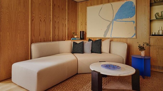 A cozy seating nook featuring a minimalist cream sectional, abstract artwork, and vibrant blue side table against wood-paneled walls.