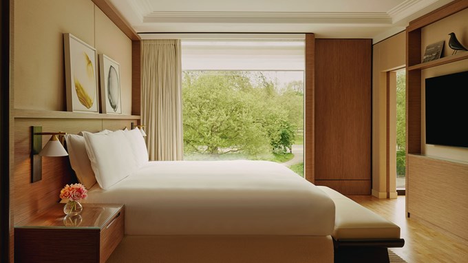 A serene bedroom with a king-sized bed, neutral decor, and large windows showcasing a tranquil green landscape.