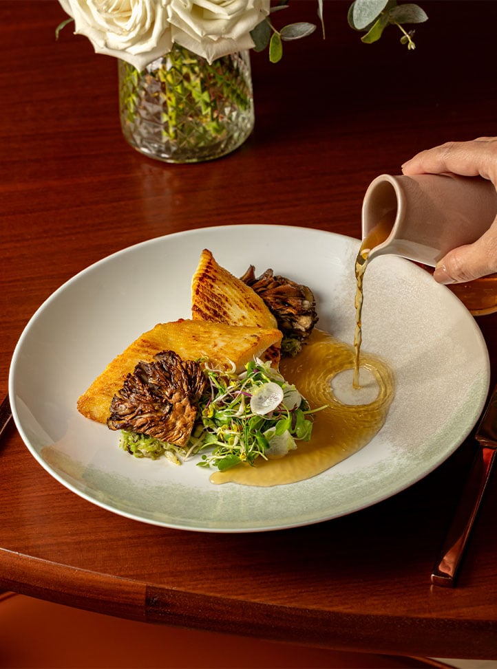 Sauce being poured over a plated dish with roasted vegetables and microgreens on a white plate.