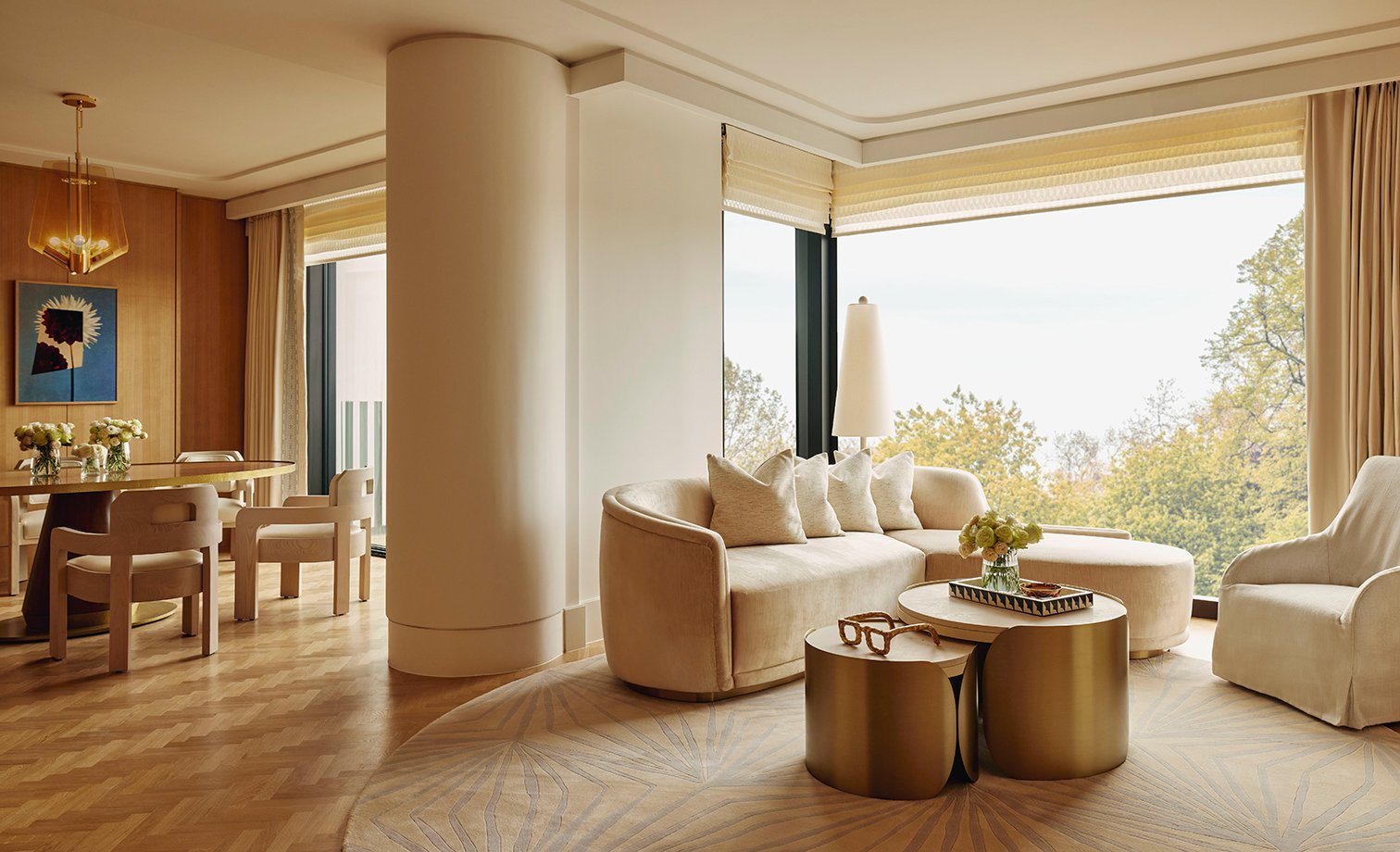 Interior of the Deluxe Balcony Park Suite. Cream sofas are set around a bronze coffee table which is topped with books and flowers. To the left is a dining table with chairs.