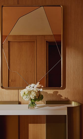 A geometric mirror on the wall, in front of it is a vase of cream flowers