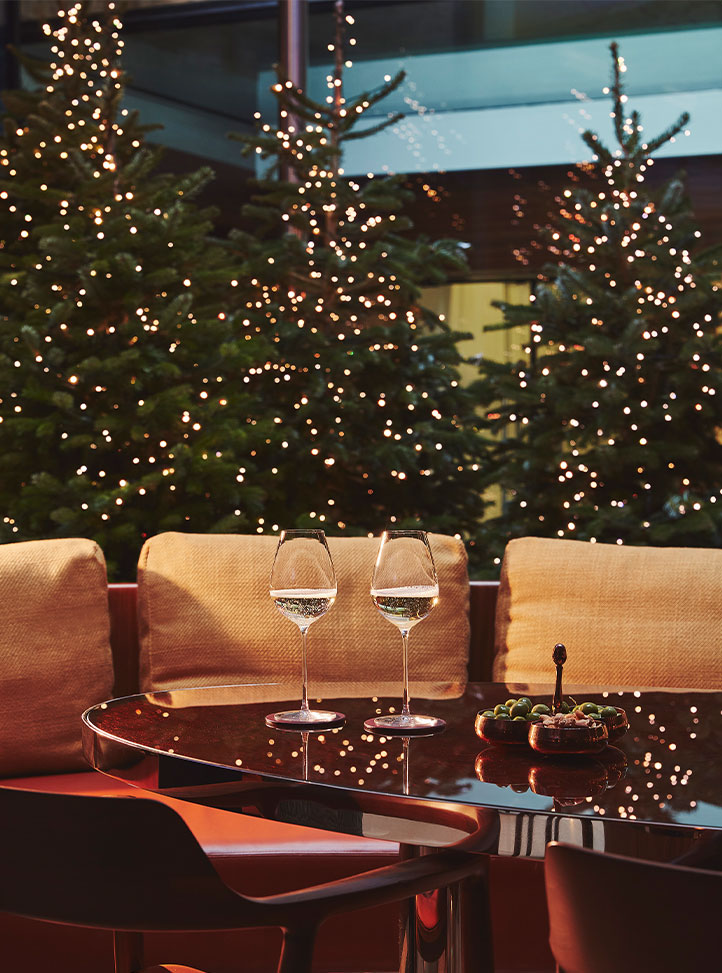 Two wine glasses and a dish of snacks rest on a glossy black table with cushioned seating, set against a backdrop of outdoor Christmas trees lit with twinkling string lights.
