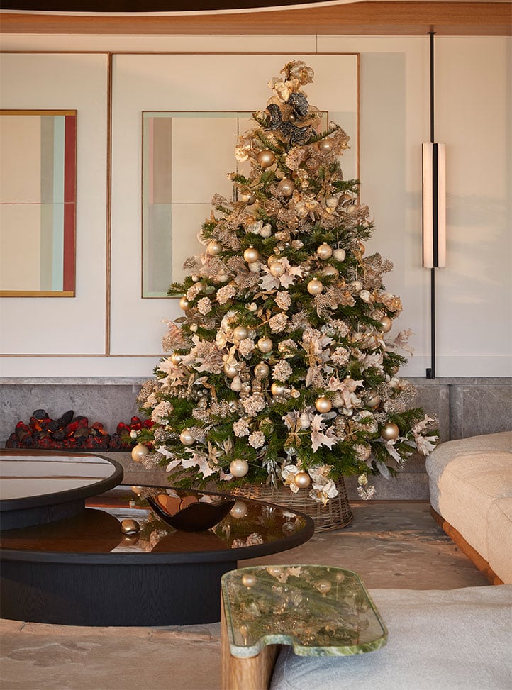 A Christmas tree decorated with gold ornaments, flowers, and ribbons stands in a modern lounge beside a low round coffee table and a cream sofa.