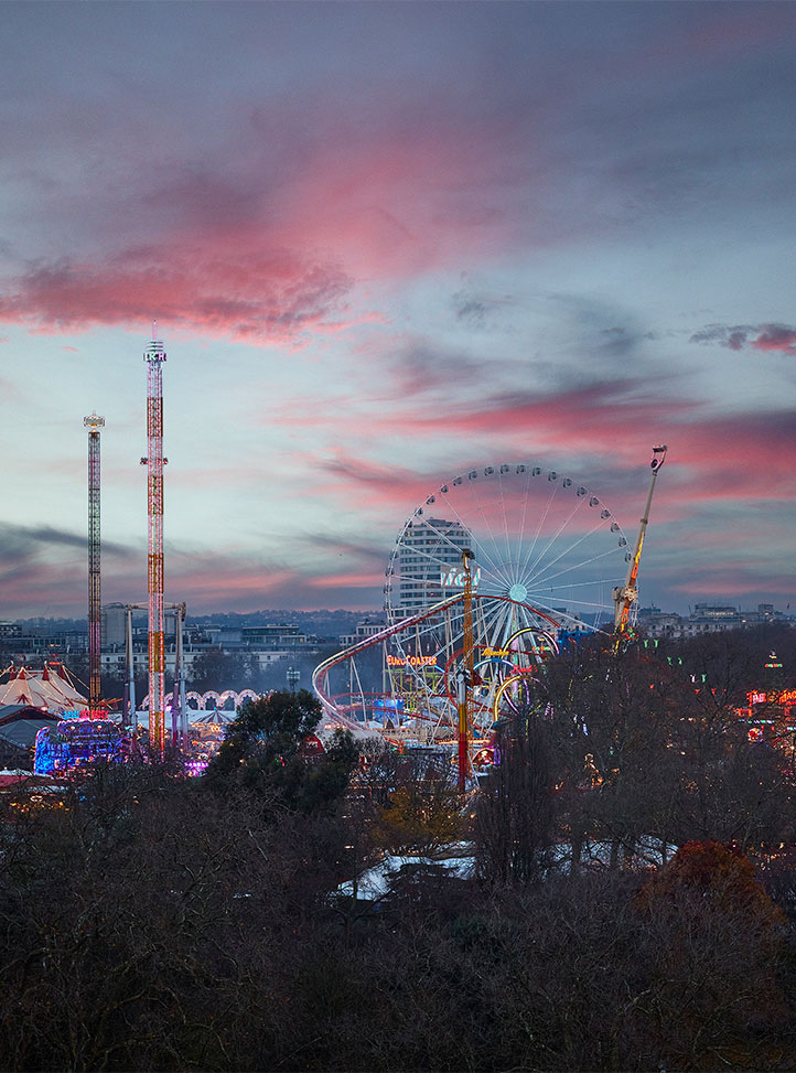 A view of a fairground at dusk with a Ferris wheel, rollercoaster, and tall rides lit up against a dramatic pink and purple sunset sky.