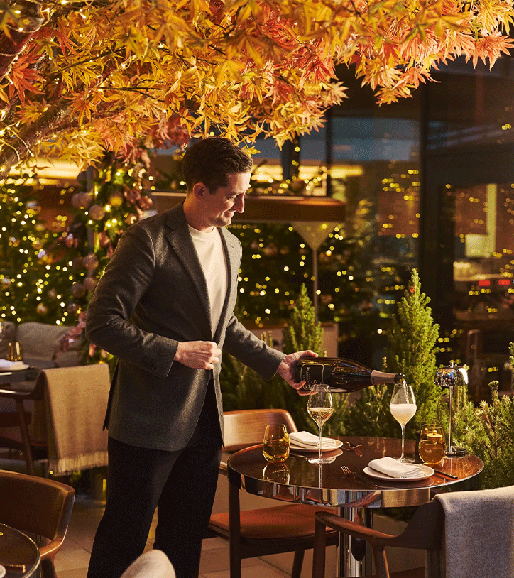 Staff member pouring sparkling wine at a festive restaurant table beneath an illuminated autumn tree and twinkling lights.
