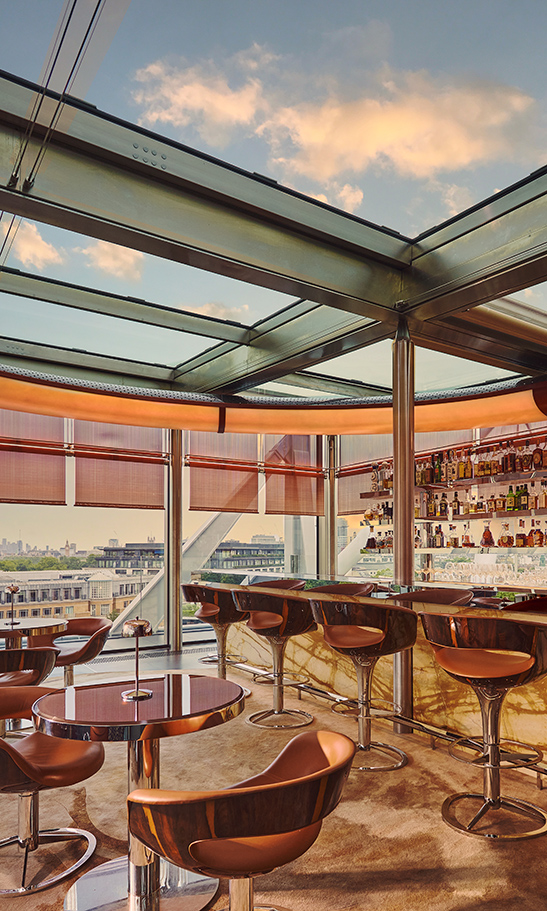 Rooftop bar with marble counter, leather stools, and round tables set beside floor-to-ceiling windows offering panoramic city views at sunset.