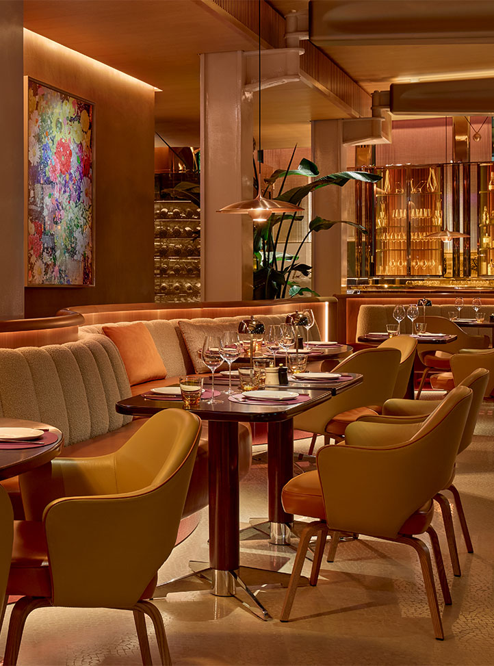 Elegant restaurant interior with curved banquette seating, mustard and terracotta chairs, and tables set with wine glasses under warm lighting.