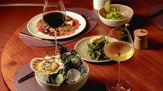 A fine dining table featuring scallop ceviche, salad, red and white wine, and beef carpaccio on dark wood.