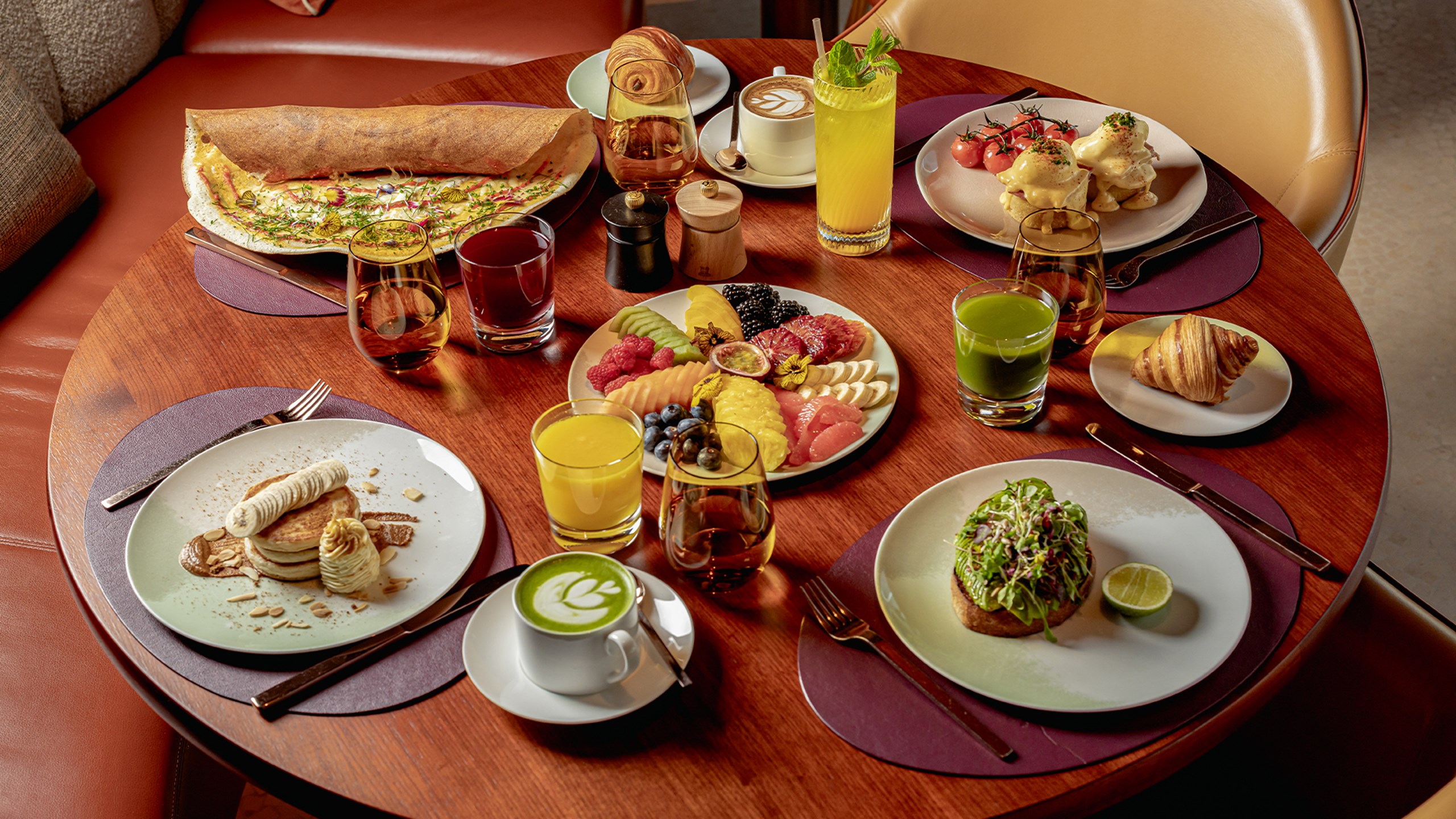 A colourful breakfast spread with pancakes, avocado toast, fresh fruit, eggs Benedict, pastries, juices, and coffee on a round table.