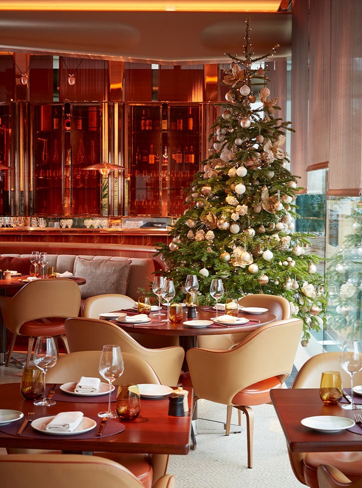 A festive dining area with round wooden tables set for service, tan leather chairs, and a large Christmas tree decorated with gold and white ornaments beside floor-to-ceiling windows.