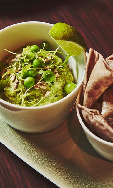 A dish of spring pea guacamole served at abc kitchens. The guacamole is green and topped with peas, to the right are pitta chips for dipping