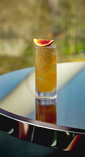 Close-up of a tall cocktail served in a ribbed glass, garnished with a slice of blood orange and topped with a fine dusting of spice. The drink is placed on a reflective outdoor table with a blurred background of greenery.