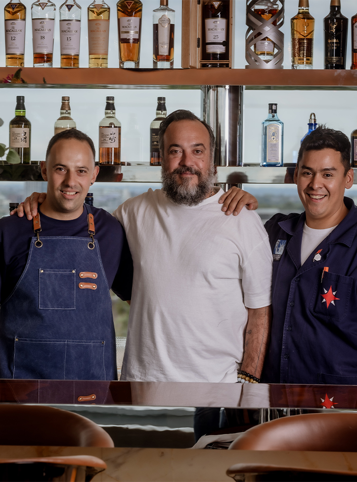 Tato smiling with a team member either side of him, in front of rows of spirits at The Emory Rooftop…