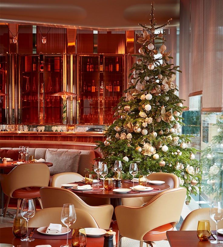 Festive dining room with a decorated Christmas tree beside neatly set tables and warm ambient lighting