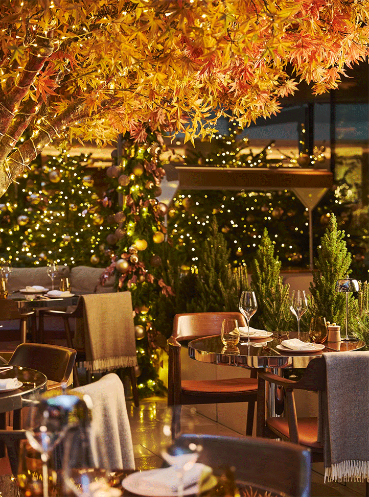 Festive restaurant terrace with autumn foliage, twinkling lights, decorated trees, and elegantly set tables at dusk.