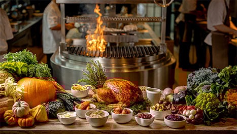 A roast turkey with bowls of sides arranged on a counter, with an open flame grill in the background