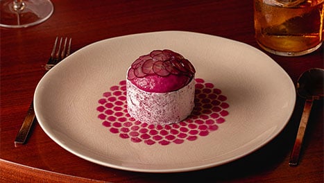 A plated purple-topped dessert on a patterned plate beside a cocktail and a floral arrangement.