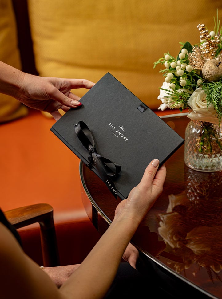 Hands hold a black The Emory gift voucher envelope beside a small festive floral arrangement on a glossy table.