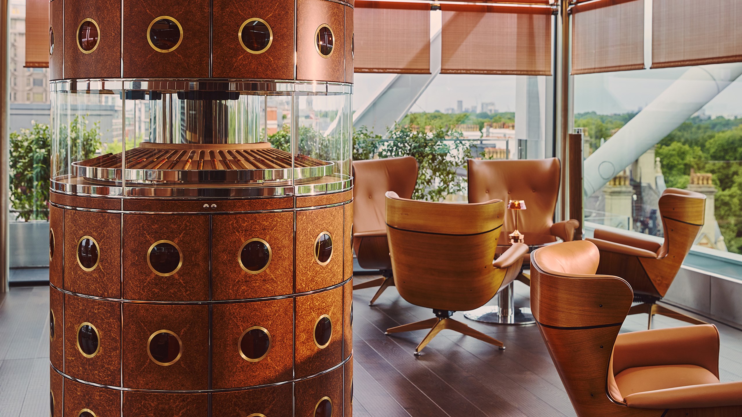 Circular wooden cigar humidor with glass display surrounded by tan leather armchairs and low tables in a modern lounge with floor-to-ceiling windows overlooking greenery.