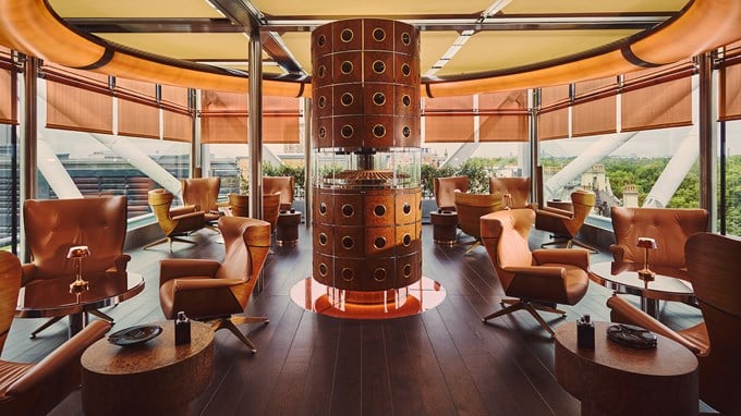 Circular wooden cigar humidor with glass display surrounded by tan leather armchairs and low tables in a modern lounge with floor-to-ceiling windows overlooking greenery.