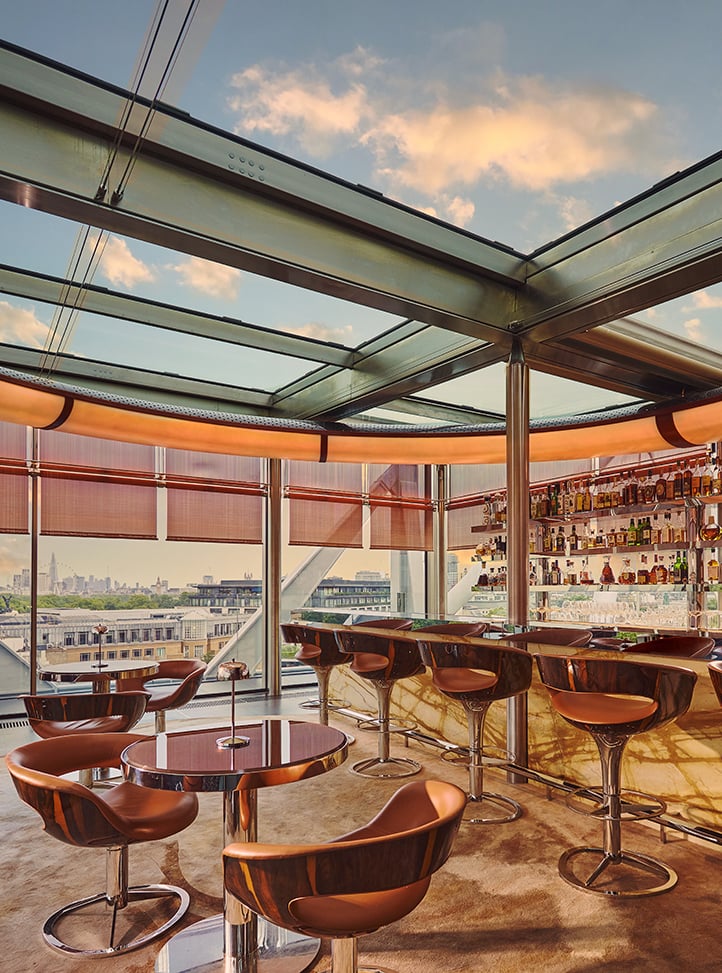 Rooftop bar with marble counter, leather stools, and round tables set beside floor-to-ceiling windows offering panoramic city views at sunset.