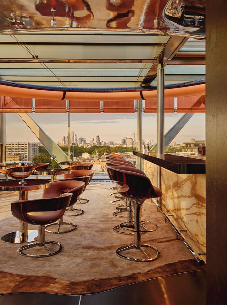 A stylish rooftop bar with caramel-toned seating, a marble counter, and panoramic views across the London skyline through floor-to-ceiling glass.