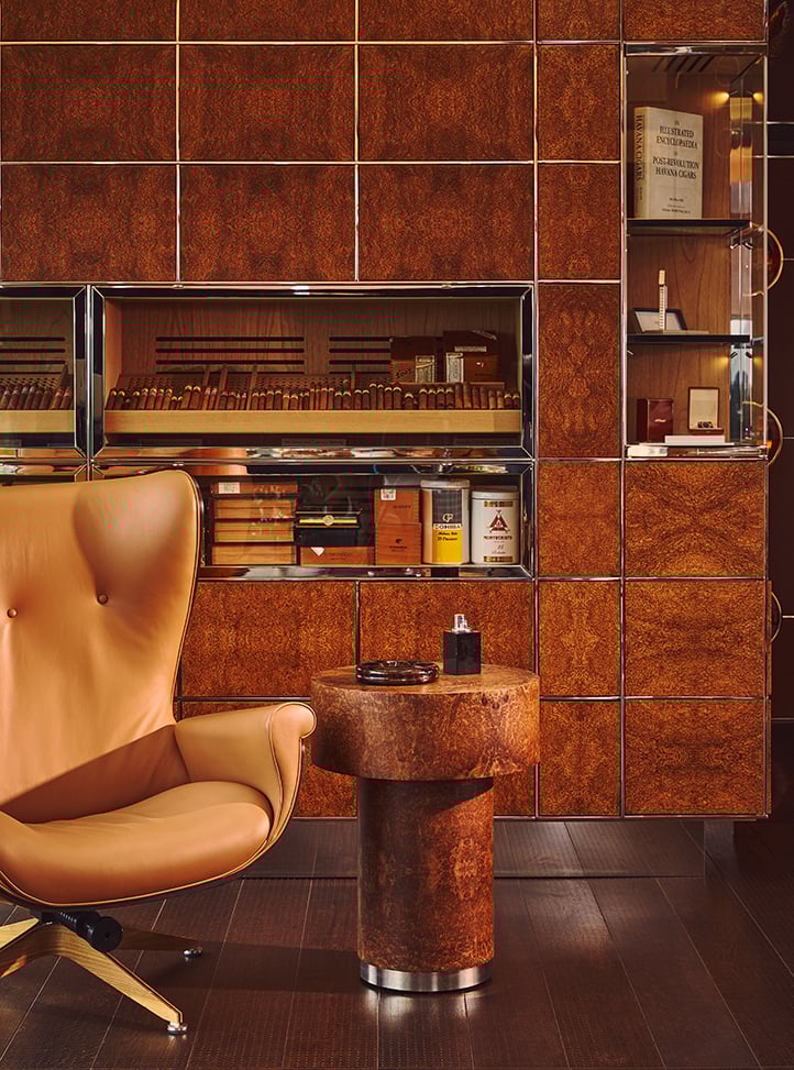 Leather armchair and round wooden side table with an ashtray in front of a cigar humidor cabinet filled with cigars and cigar boxes in a luxurious lounge setting.