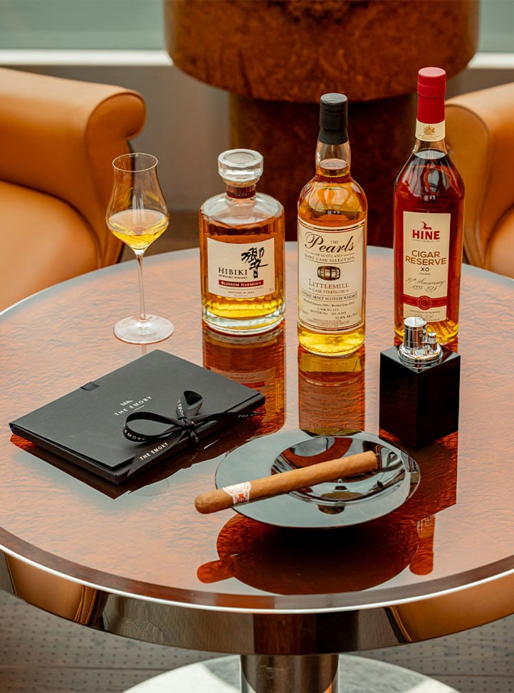 Three bottles of whisky, a cigar on an ashtray, and a notepad on a polished round table between two leather armchairs.