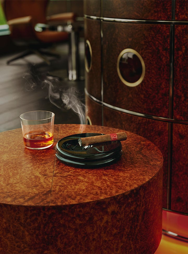 A close-up of a lit cigar resting in a black ashtray beside a glass of whisky on a polished, burl wood side table in a refined lounge setting.