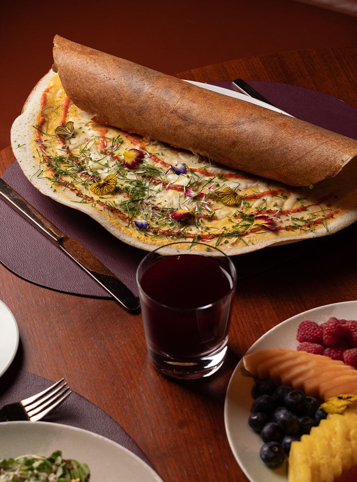 A colourful dosa topped with herbs and edible flowers is partially rolled and served with a dark red juice and fresh fruit on the side.