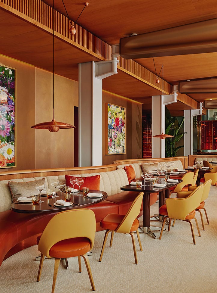 A warm, contemporary restaurant interior with curved banquettes, mustard and rust chairs, and vibrant floral artwork on the walls.
