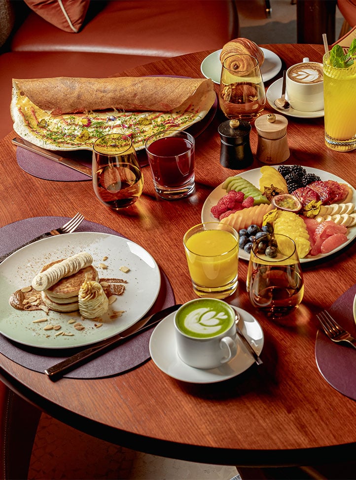 A luxurious brunch spread on a round wooden table featuring pancakes with banana, a floral dosa, fresh tropical fruits, juices, a croissant, and a matcha latte with latte art.