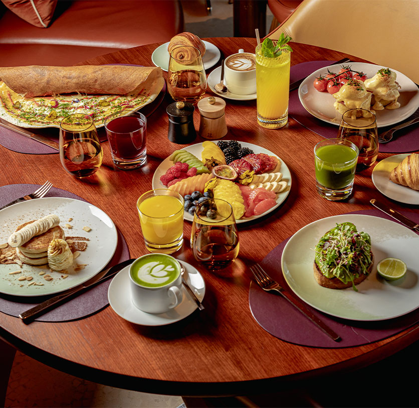 A bountiful brunch spread featuring pancakes, avocado toast, croissants, juices, colourful fruits, a dosa, and cappuccinos on a round wooden table.