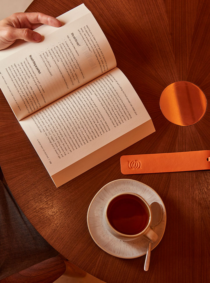 A person reads an open book at a circular wooden table with a cup of tea, spoon on the saucer, and an orange leather bookmark embossed with a wave-like logo resting nearby.
