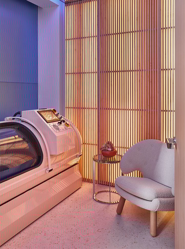 A private wellness suite featuring a sleek white hyperbaric oxygen therapy chamber, a curved boucle armchair, and a softly lit slatted wood wall creating a serene, modern ambience.