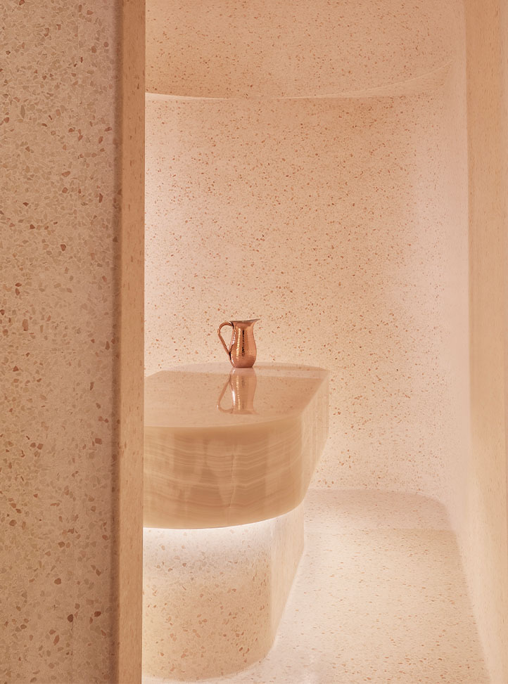 A softly lit, minimalist hammam space with terrazzo walls and flooring, featuring a central onyx treatment table topped with a copper pitcher for traditional cleansing rituals.