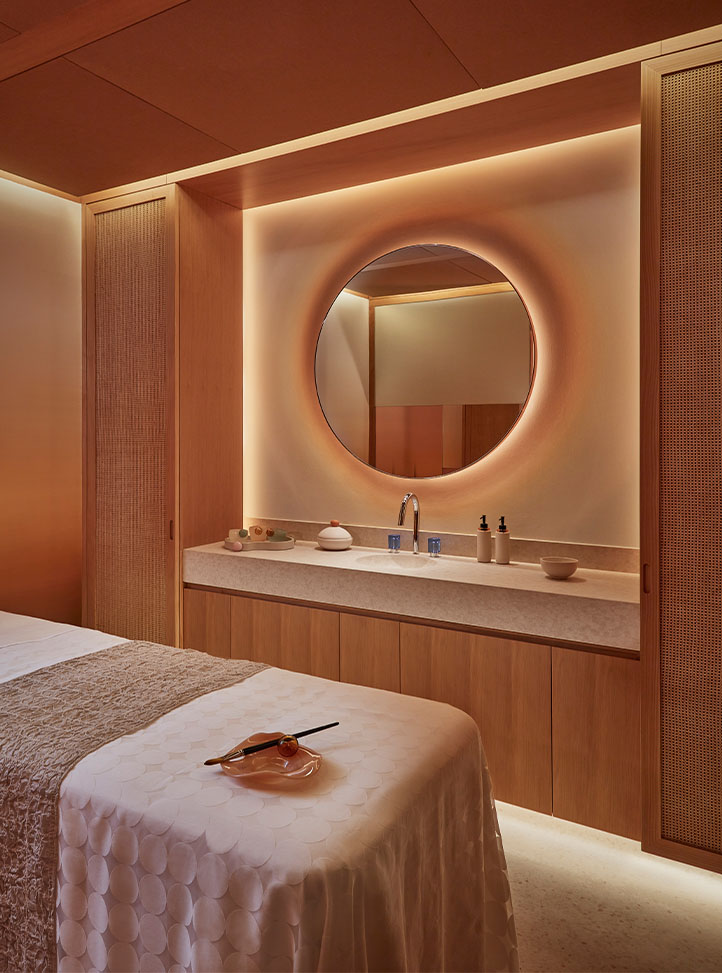 A warmly lit spa treatment room with a massage bed covered in a textured white throw, wooden cabinetry, and a round backlit mirror above a countertop with ceramic spa accessories.