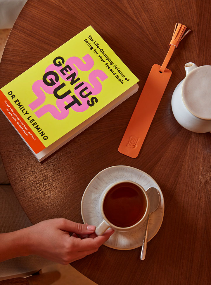 A close-up of a wooden table with a hand holding a cup of tea, a bright yellow wellness book titled 