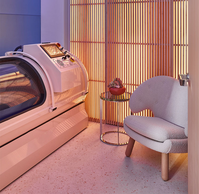 A private wellness suite featuring a sleek white hyperbaric oxygen therapy chamber, a curved boucle armchair, and a softly lit slatted wood wall creating a serene, modern ambience.