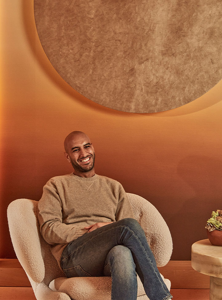 A smiling man sits relaxed in a cream textured armchair, wearing a beige jumper and jeans, with a warm-toned circular wall art and gradient orange background behind him.