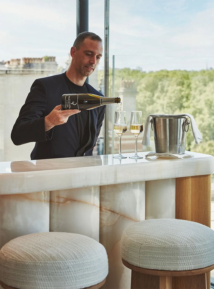 Man pours champagne into flutes at a marble bar with stools and a view of trees through large windows.