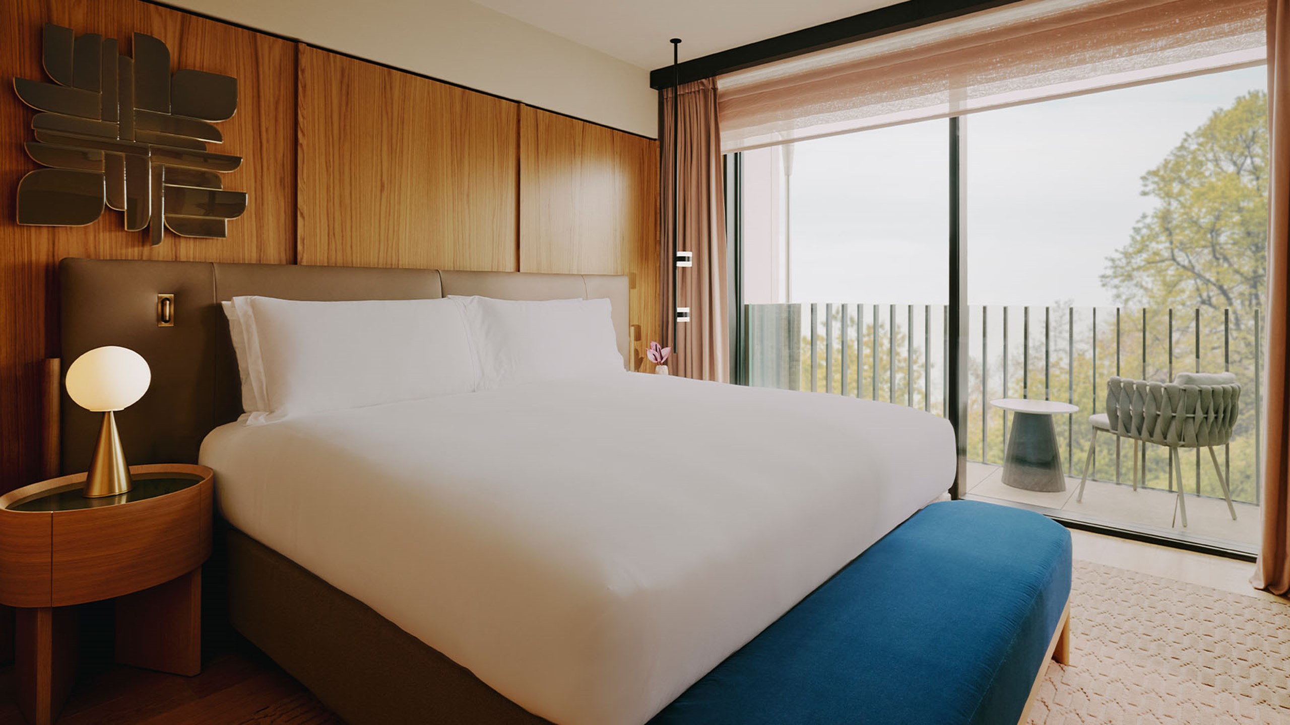 Balcony Park Suite bedroom. A double bed sits in the foreground and a bench sits at the end of the bed. A round wooden bedside table and lamp sits beside it. A large window sits behind the bed.
