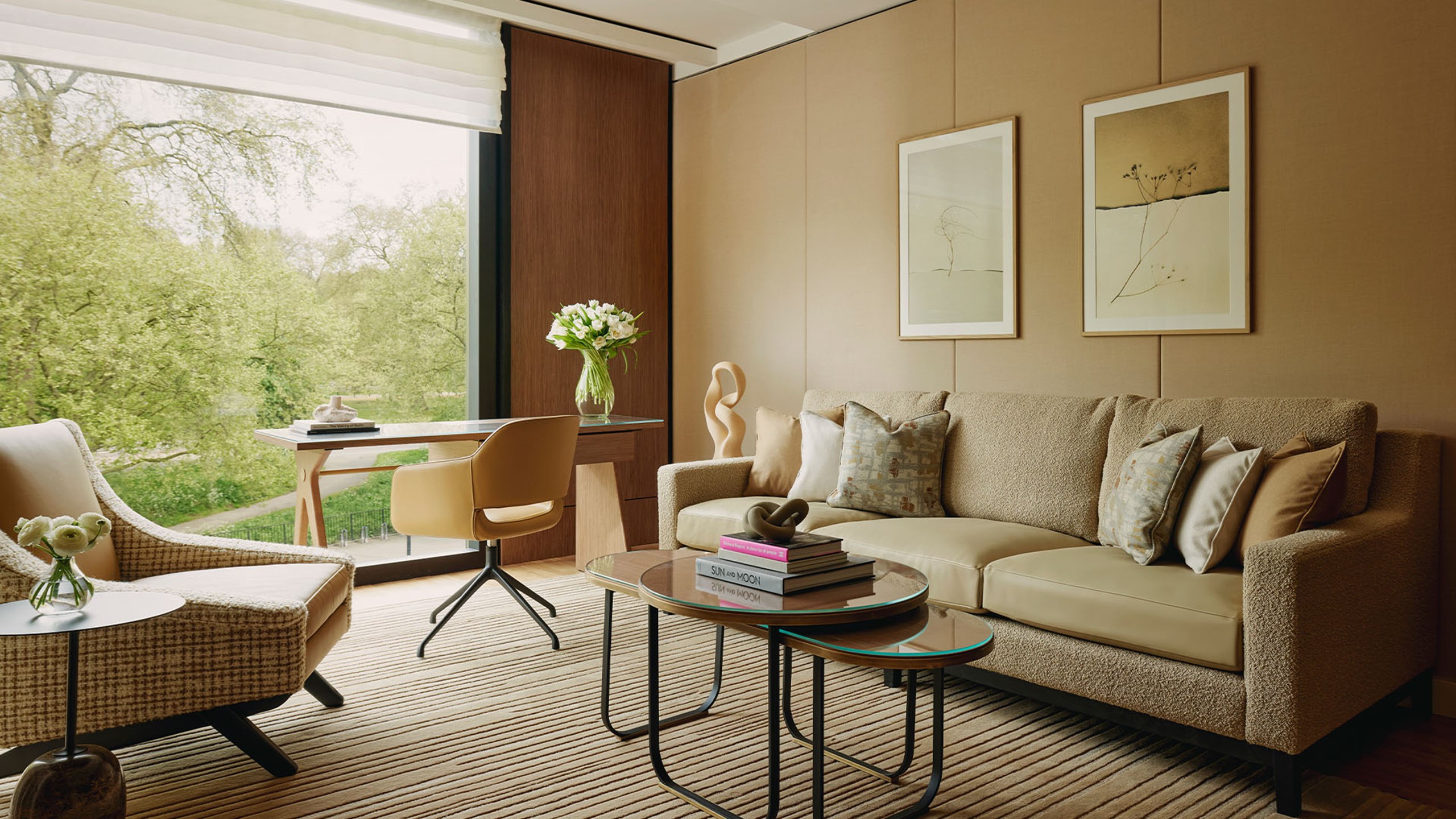 Elegant living area with a neutral-toned sofa, abstract artwork, coffee table, desk, and armchair, overlooking lush greenery through a large window.