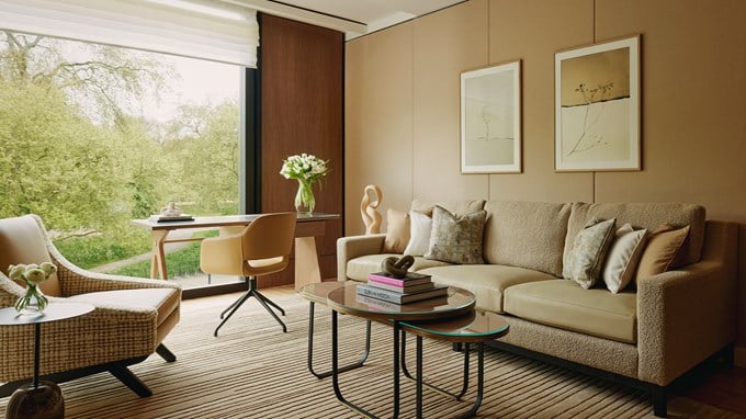 Elegant living area with a neutral-toned sofa, abstract artwork, coffee table, desk, and armchair, overlooking lush greenery through a large window.