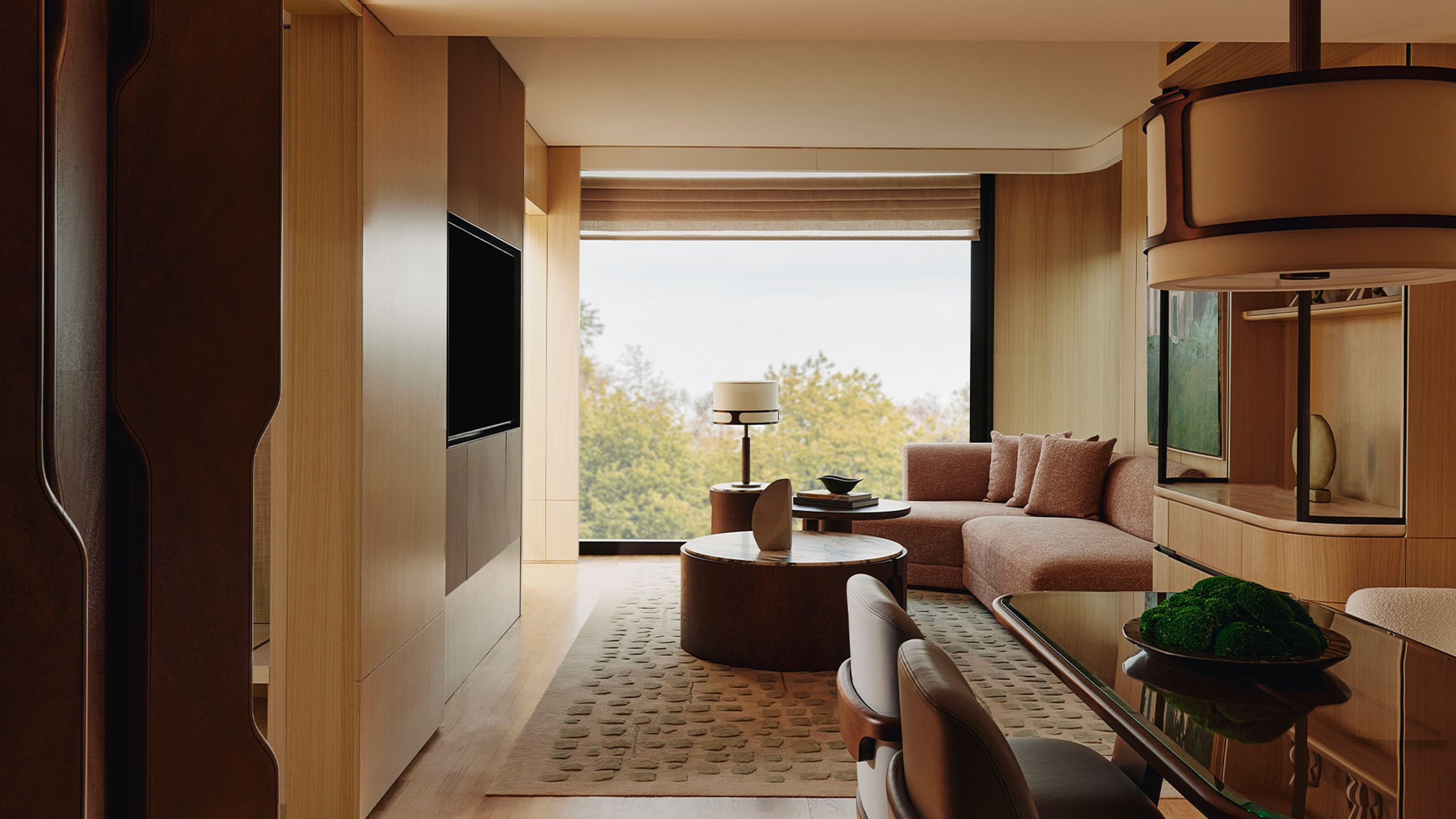 Corner Park Suite sitting area. A circular glass table and two chairs sits in the foreground. A large window sits in the background and a couch sits in front of it. A round gold and marble table with a book and a vase with flowers sits in front of the couch.