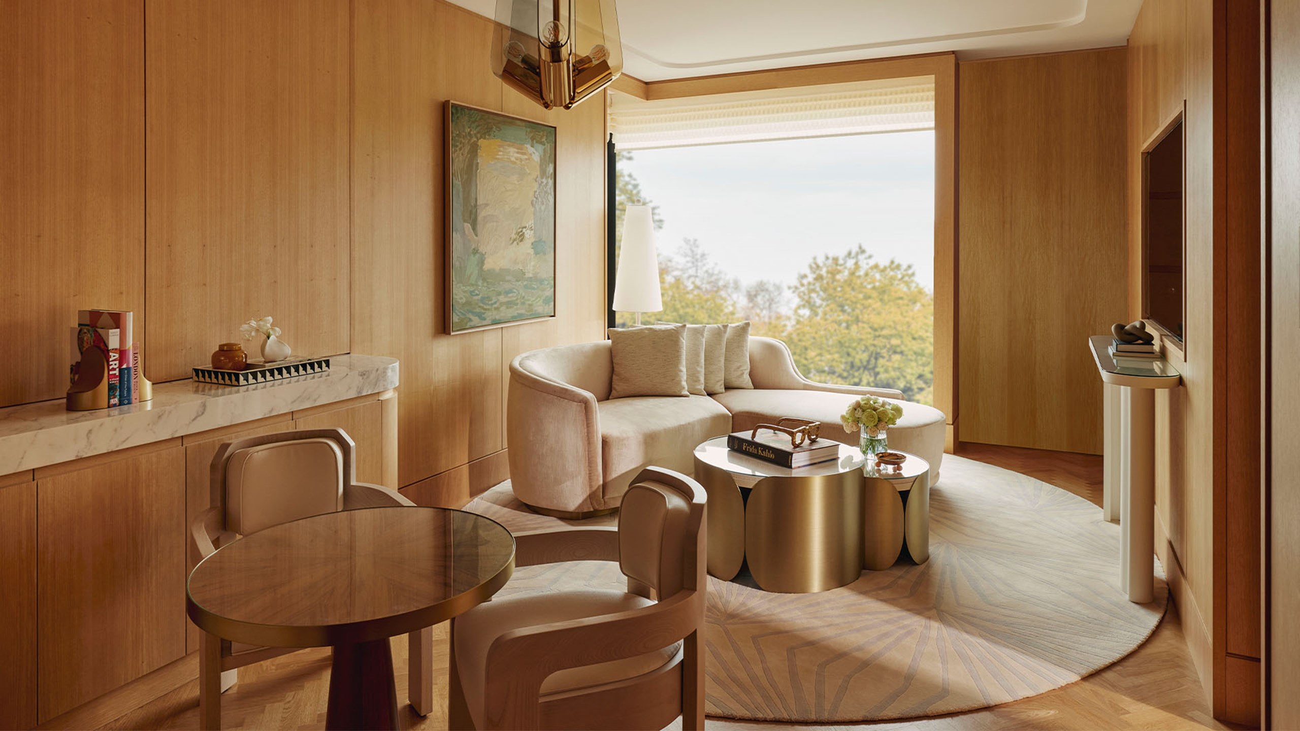 Corner Park Suite sitting area. A circular glass table and two chairs sits in the foreground. A large window sits in the background and a couch sits in front of it. A round gold and marble table with a book and a vase with flowers sits in front of the couch.