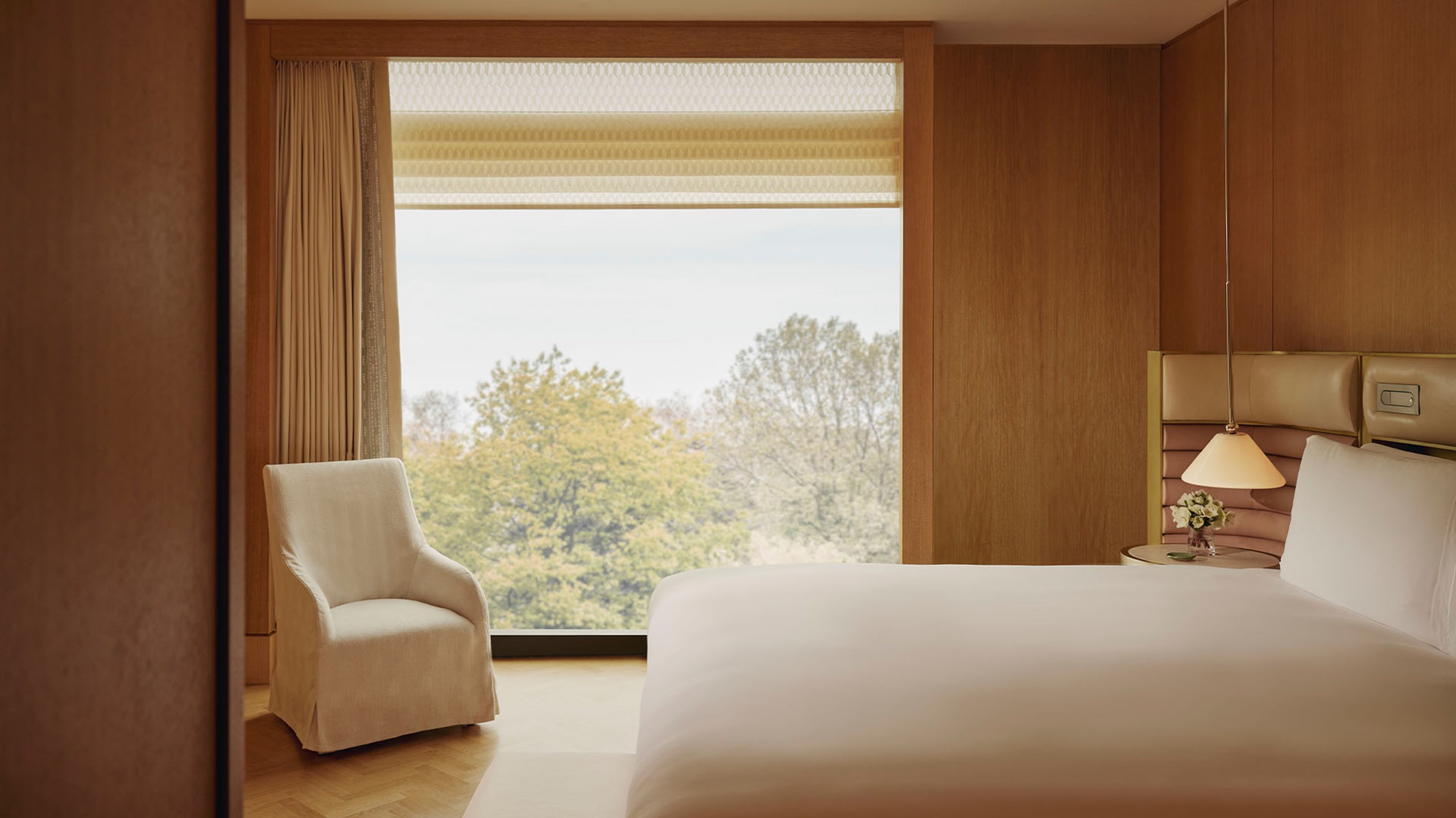 Bedroom of the Corner Park Suite with white bedding on the bed, a cream armchair in the corner and glass windows revealing the leafy green trees outside