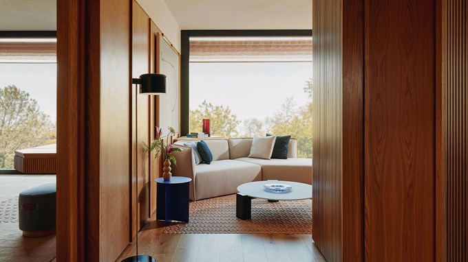 Cream sofa with dark blue cushions, placed in the sitting room of the Corner Park Suite, with large glass windows behind revealing leafy green trees